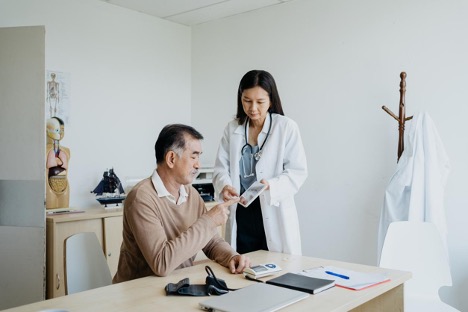 Inside the clinic, a female doctor discusses medical test results with a male patient, who points his hand at the digital tablet and asks the doctor why it is important to get routine colonoscopies.